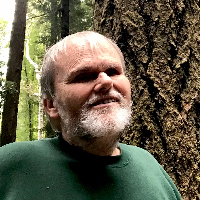 Len, smiling slightly (with a trimmed beard), stands in front of a large tree, with many trees in the background.